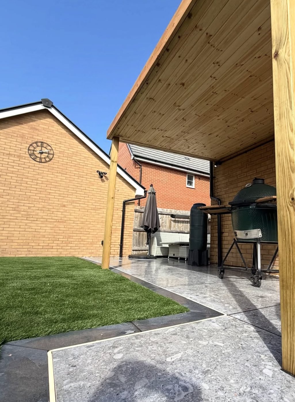 Timber canopy over a paved outdoor kitchen area with a ceramic barbecue beside an artificial grass lawn