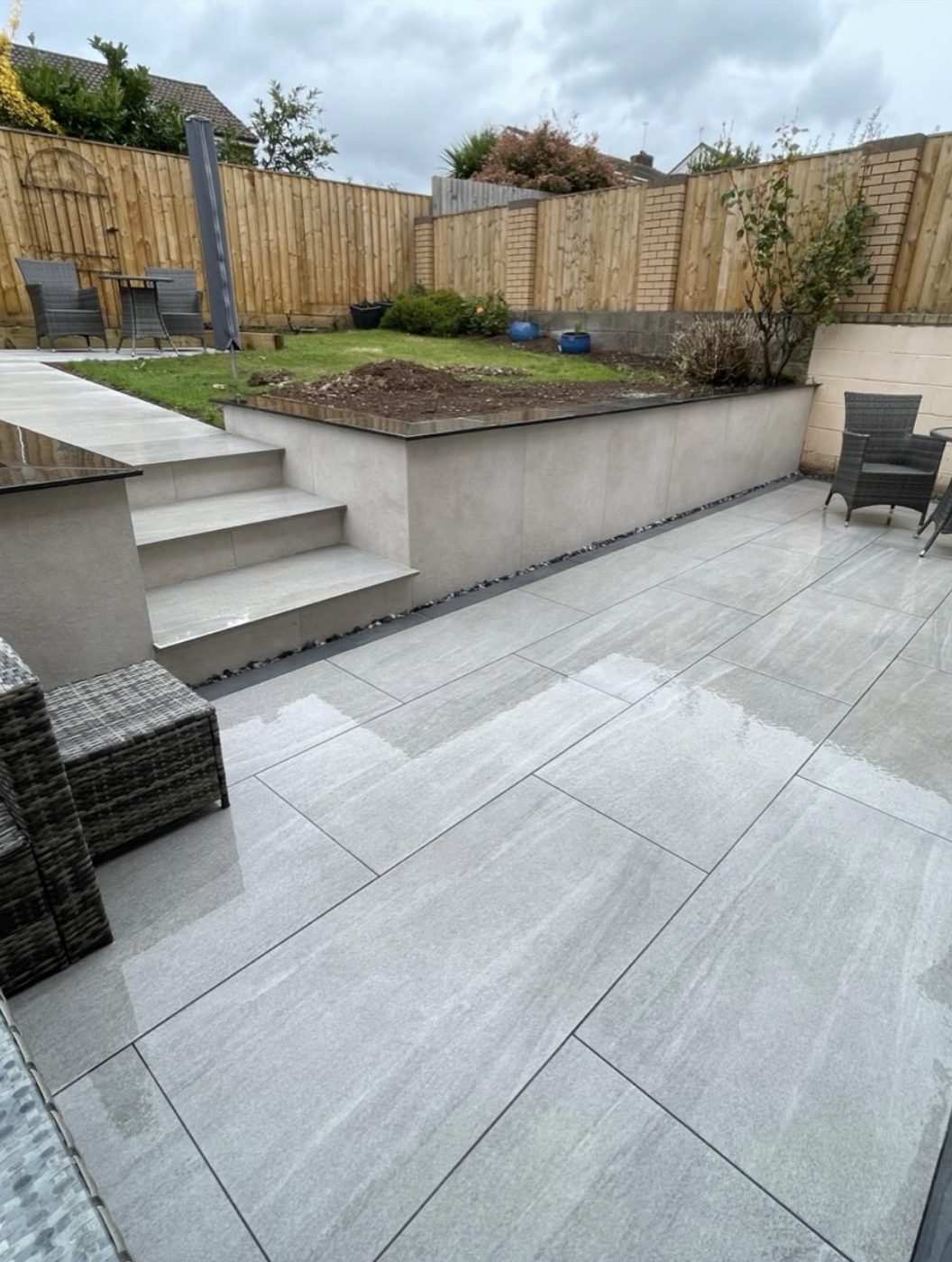 Grey porcelain patio with wide steps up to a rendered retaining wall and a raised lawn